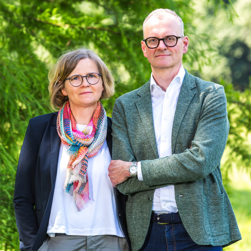Portrait of a man and woman wearing glasses standing outdoors against green foliage.