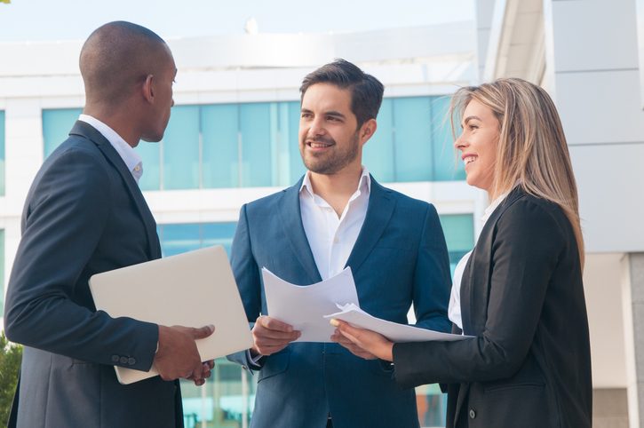 Three business professionals in suits discussing documents outdoors near an office building.