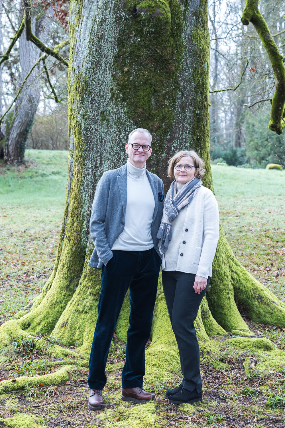 Middle-aged man and woman wearing glasses, standing side by side in front of a large moss-covered tree in a forest.