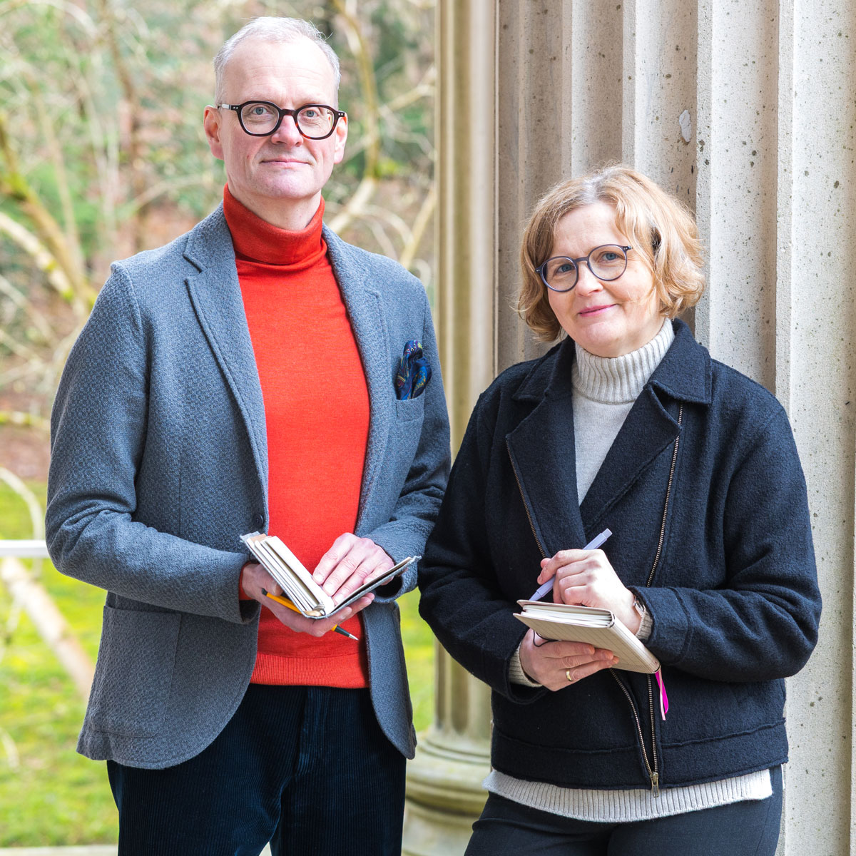 Man in gray blazer and orange turtleneck and woman in black jacket with glasses holding notebooks outdoors.