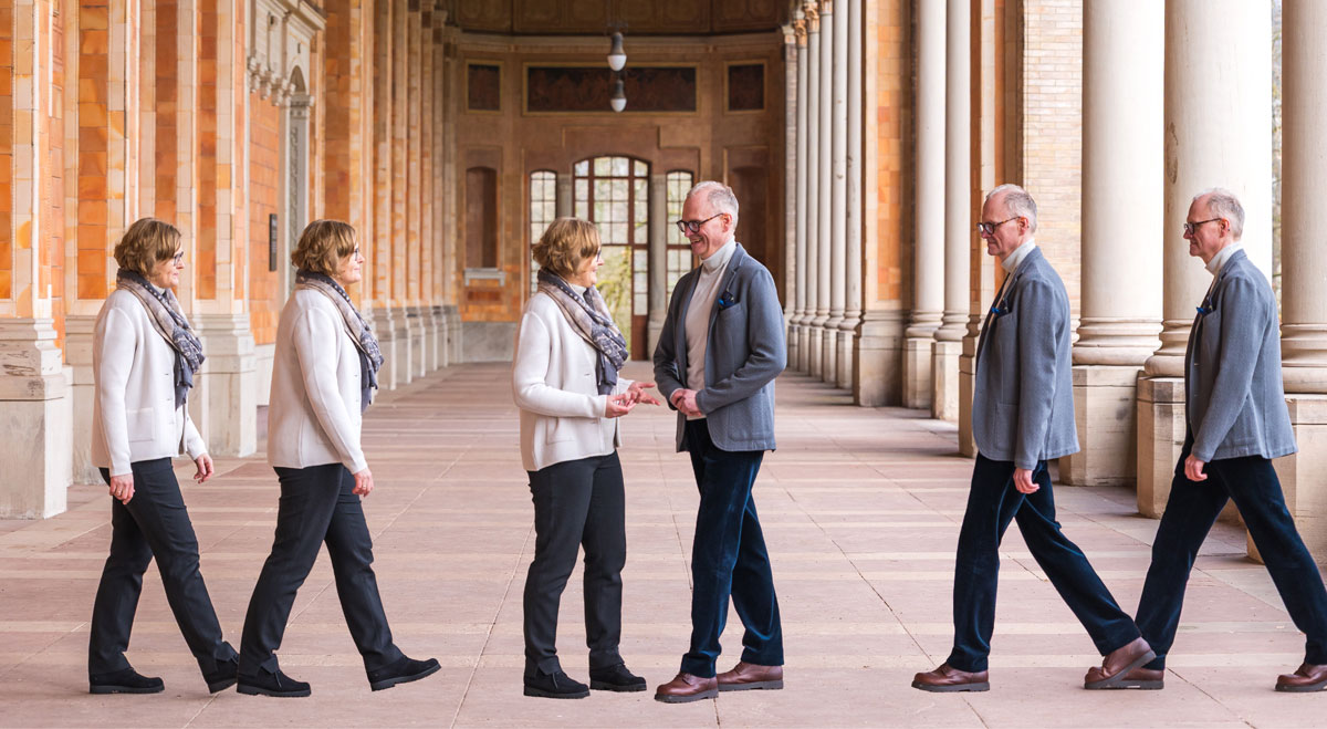 A woman and a man walk towards each other in a grand corridor and then meet and smile in the middle.