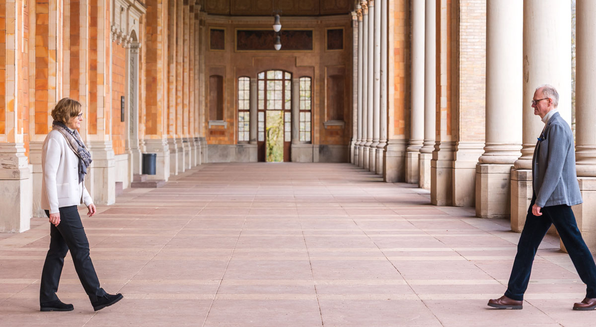 An elderly man and woman walking towards each other in a large corridor with orange brick walls and white columns.