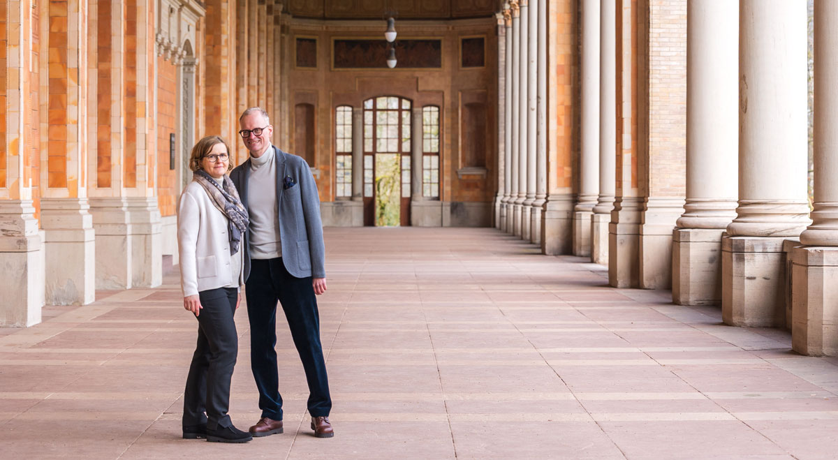 Smiling middle-aged couple standing under a columned archway with patterned walls and large windows.