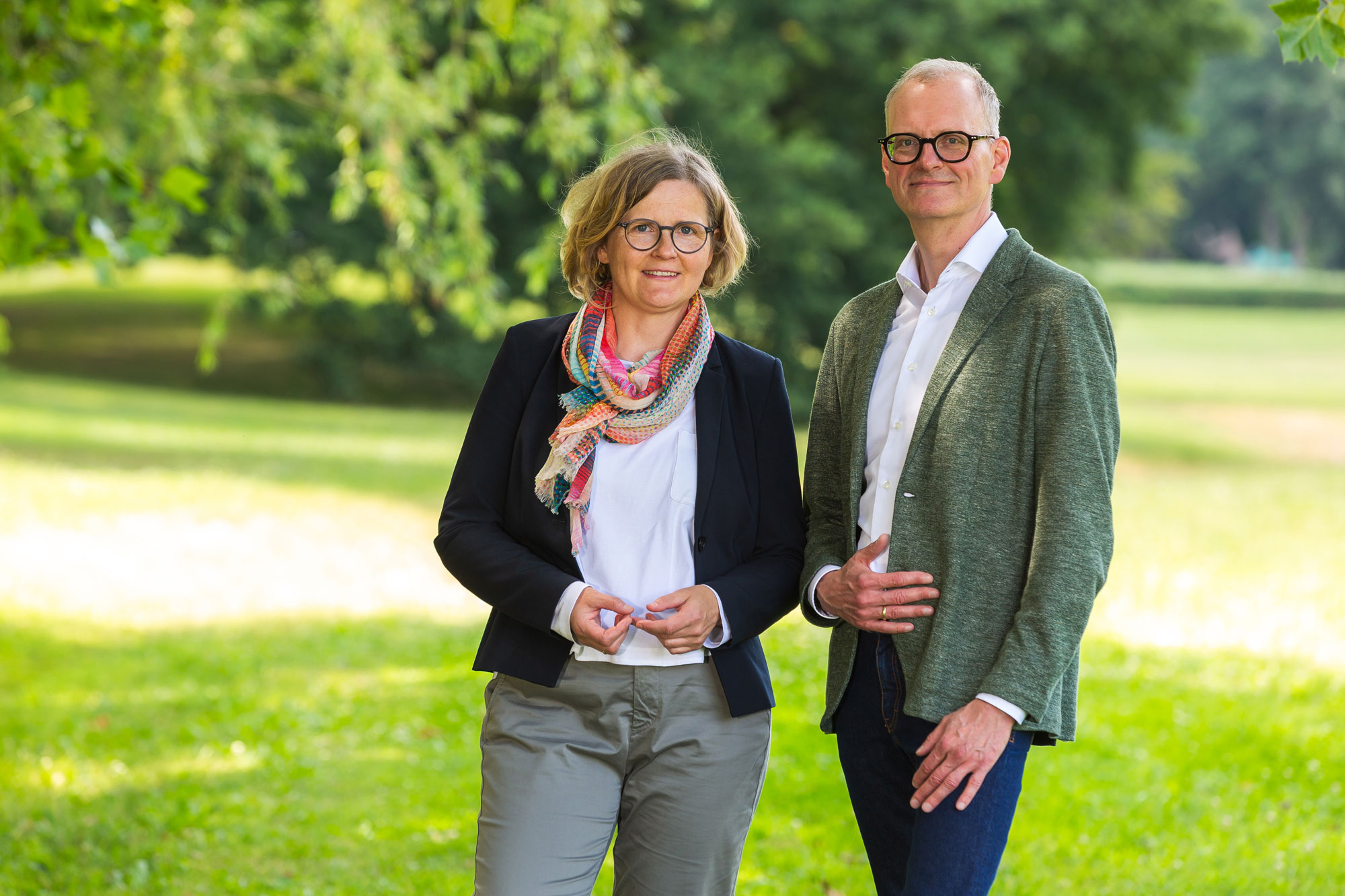 A man and woman wearing glasses stand together in a green park with trees in the background.