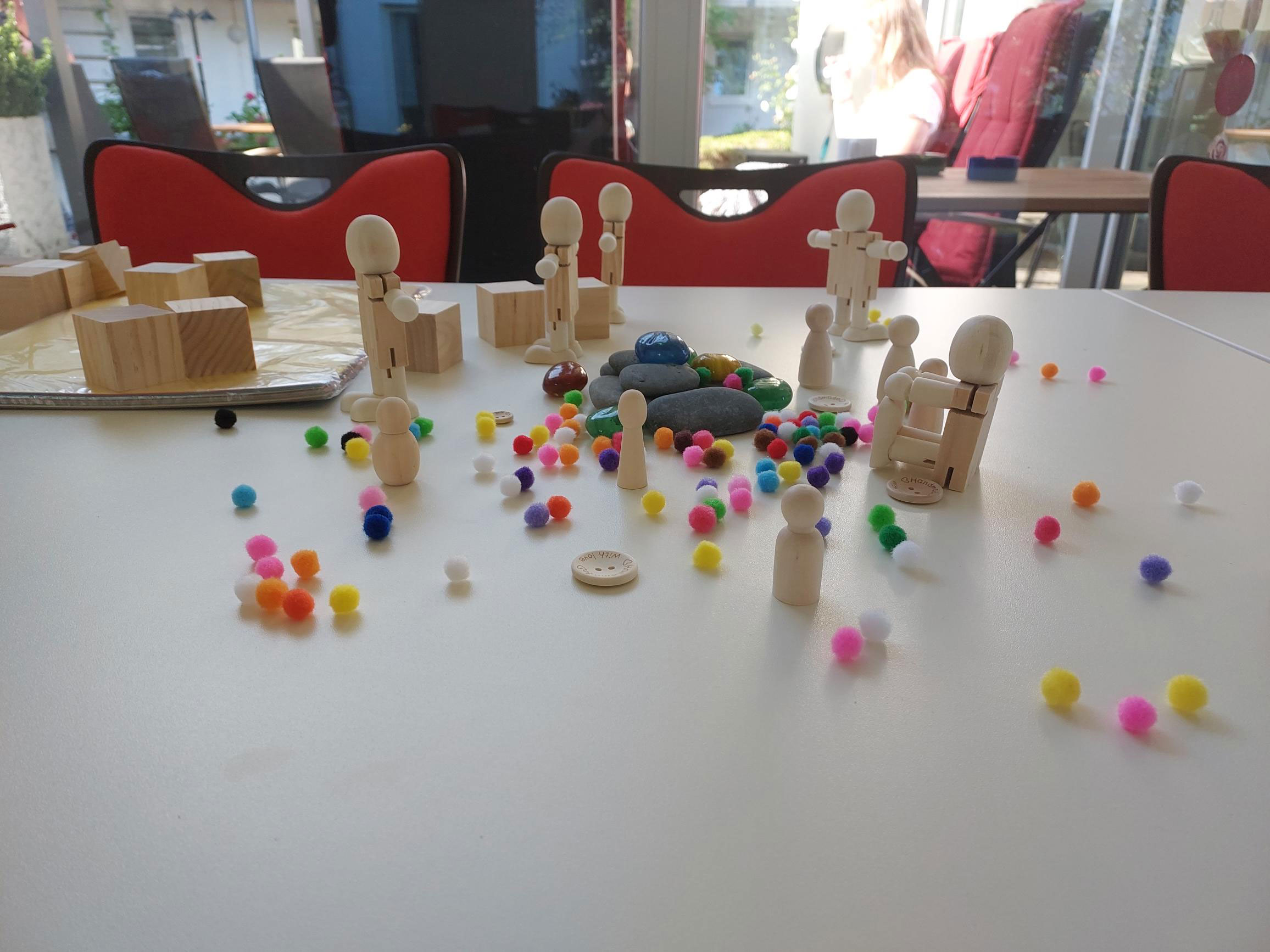 Wooden peg figures and colorful pom-poms scattered on a white table with red chairs in the background.