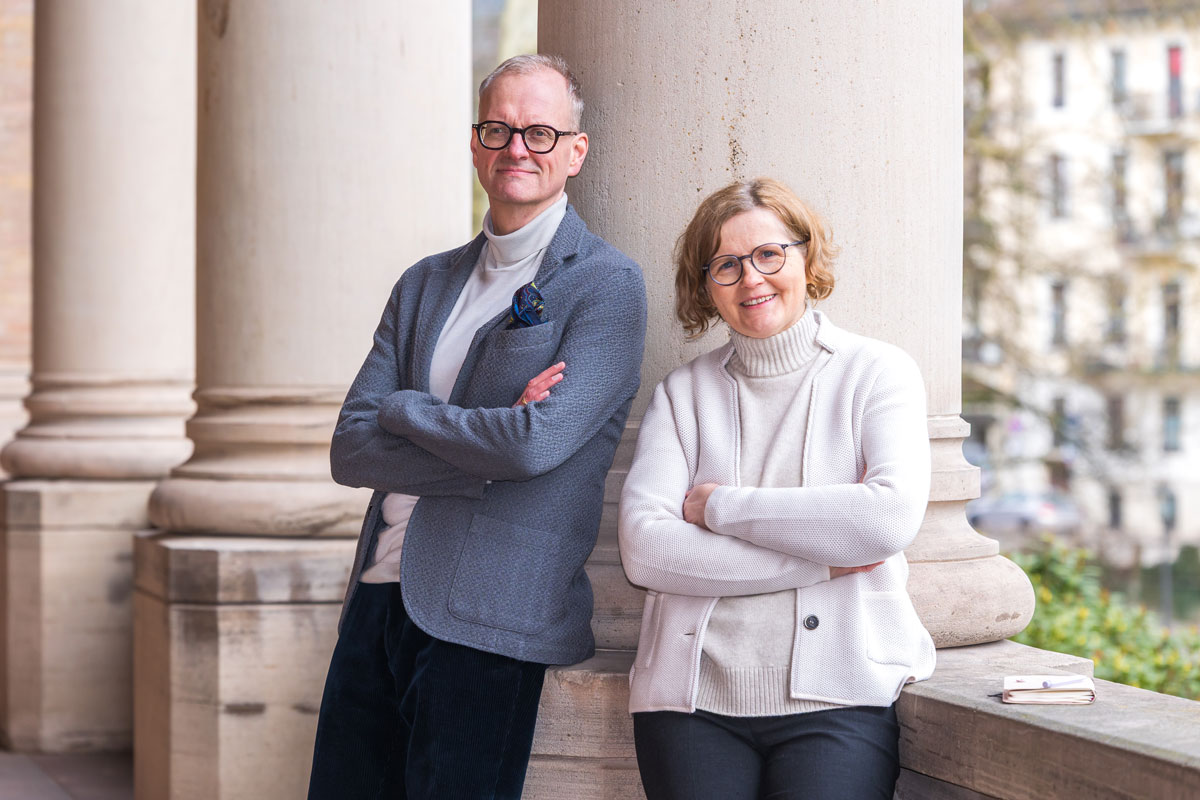 Man and woman with glasses standing with arms crossed, leaning against stone columns outdoors.
