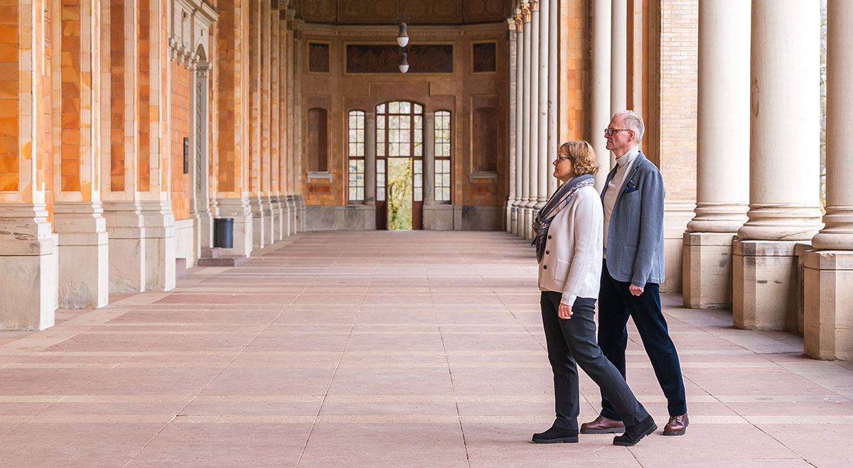 An elderly couple walking in a columned corridor of a historic building with large windows and orange brick walls.