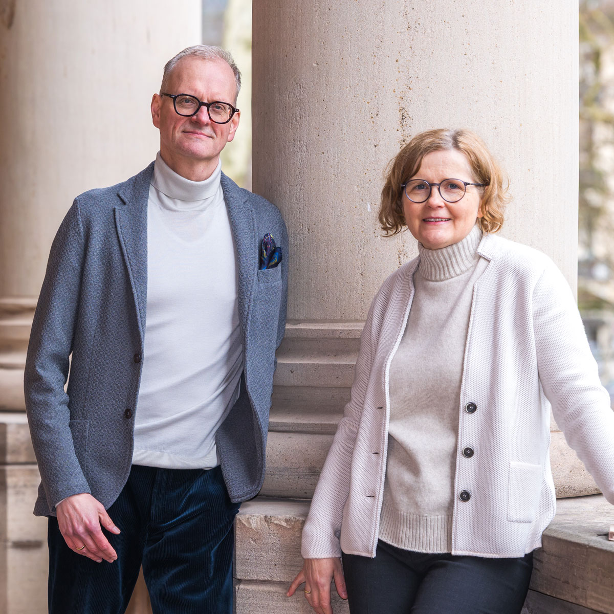 Man and woman wearing glasses and sweaters posing by stone columns outdoors.