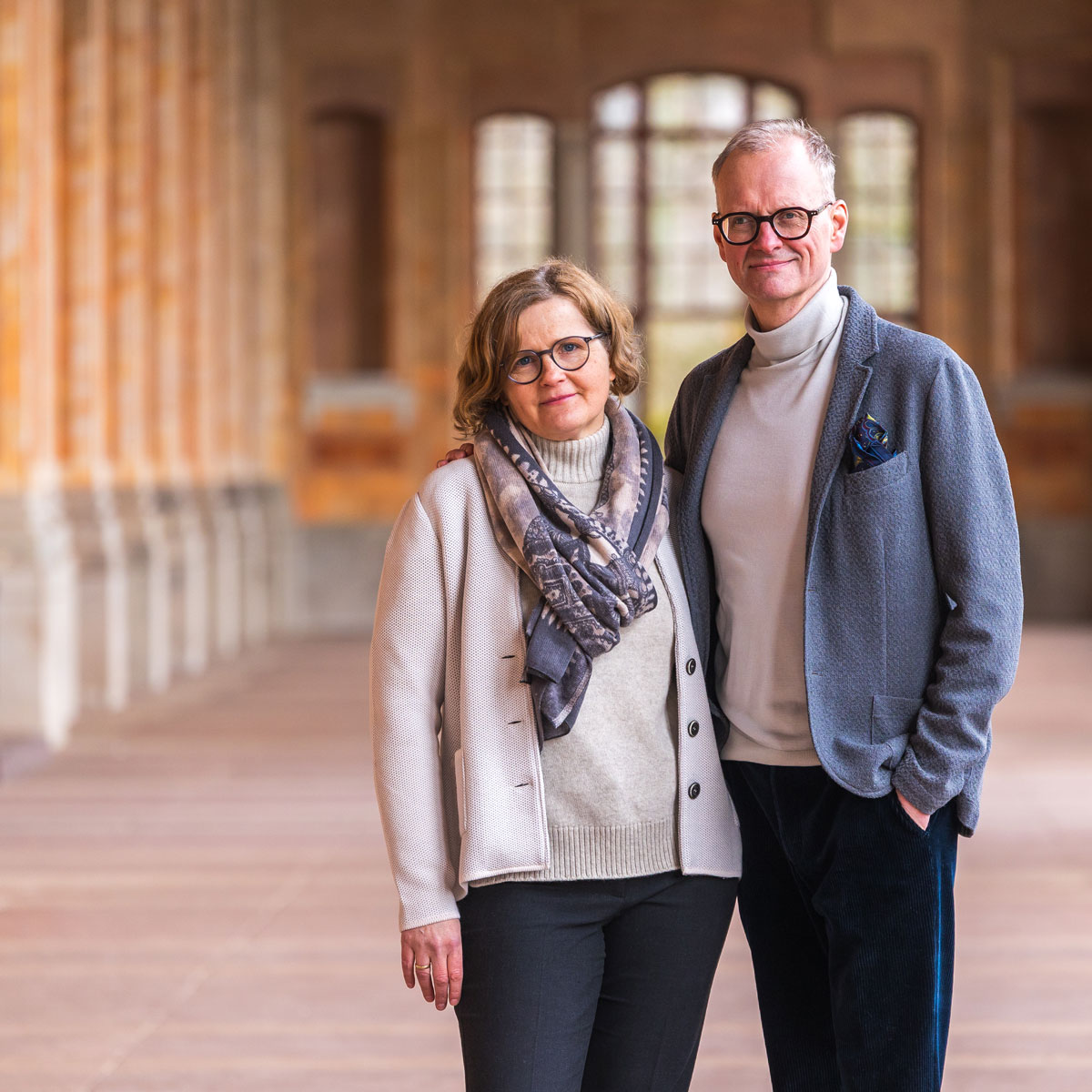 Middle-aged man and woman wearing glasses standing close together in a hallway with arches.