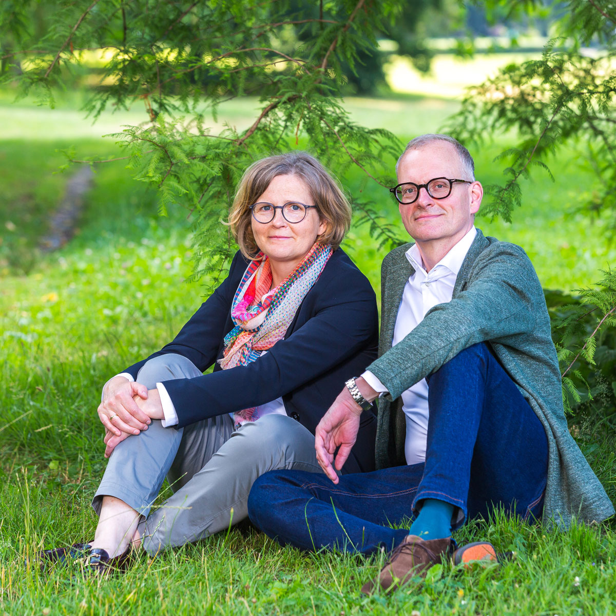 Man and woman sitting on grass outdoors under tree branches, both wearing glasses and casual clothes.