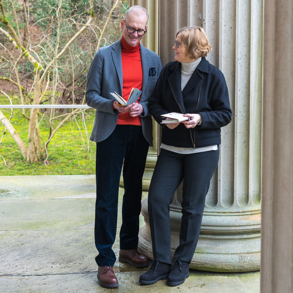 Two adults wearing glasses stand outside near a large stone column holding notebooks and smiling.