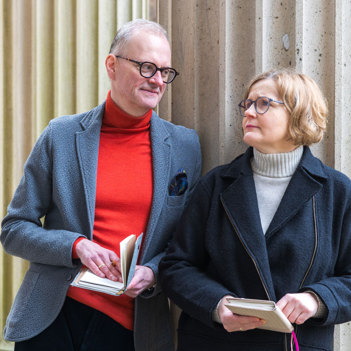 A man in glasses and a red turtleneck sweater talks with a woman in glasses and a black coat, both holding notebooks.