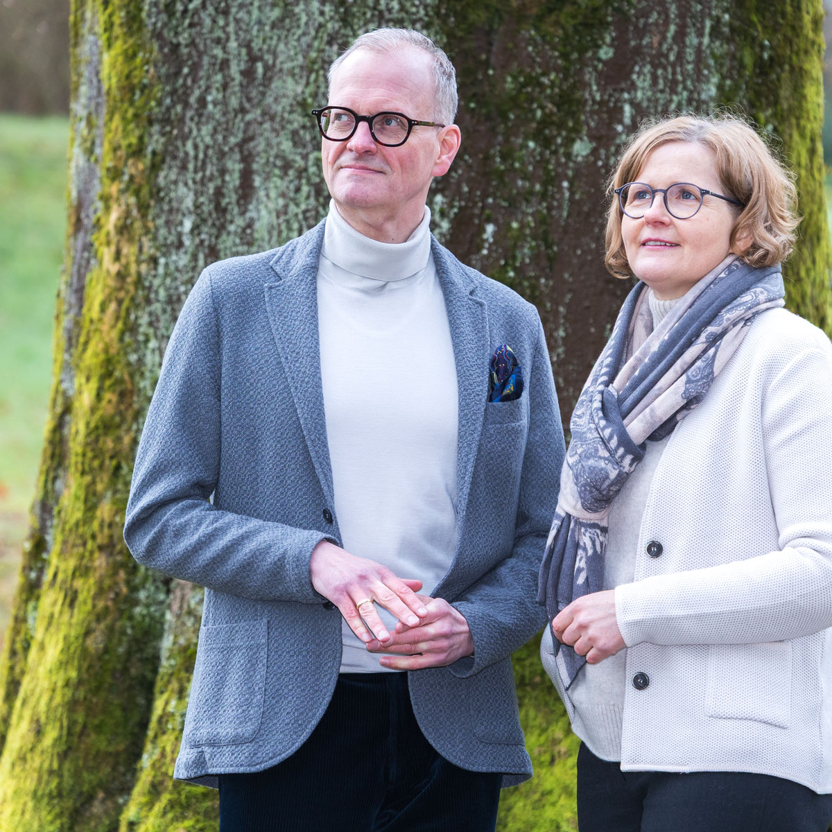 Man and woman wearing glasses standing outdoors near a large tree with moss, looking up.