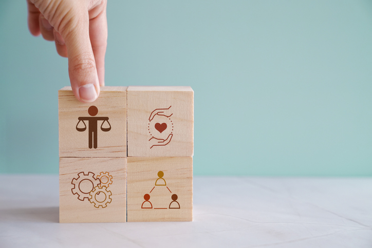 Hand placing wooden block with justice scale icon above blocks with heart, gears, and teamwork icons.