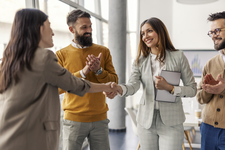 Two businesswomen shaking hands while two colleagues applaud in a modern office.