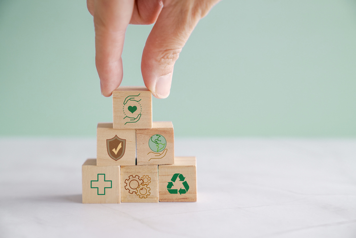Hand stacking wooden blocks with icons of health, environment, and sustainability in a pyramid.