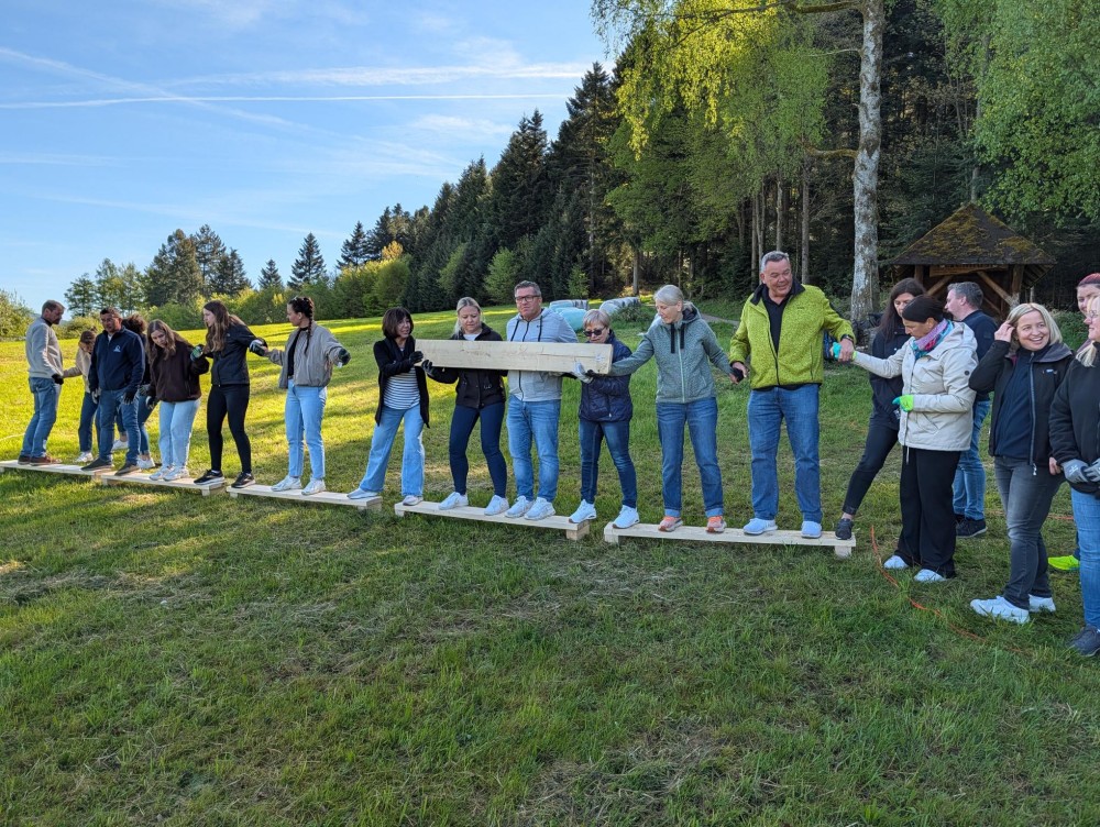 Group of people balancing on wooden planks outdoors, holding hands and supporting each other.