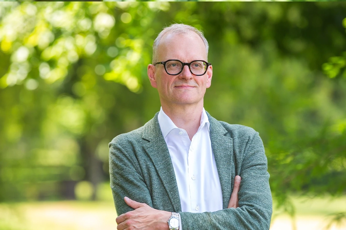 Middle-aged man wearing glasses and a gray blazer standing outdoors with arms crossed, surrounded by greenery.