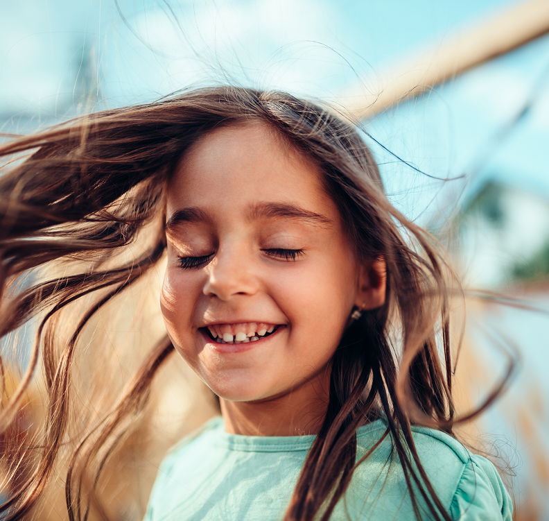 En ung jente med langt brunt hår smiler med lukkede øyne mens håret blåser i vinden.