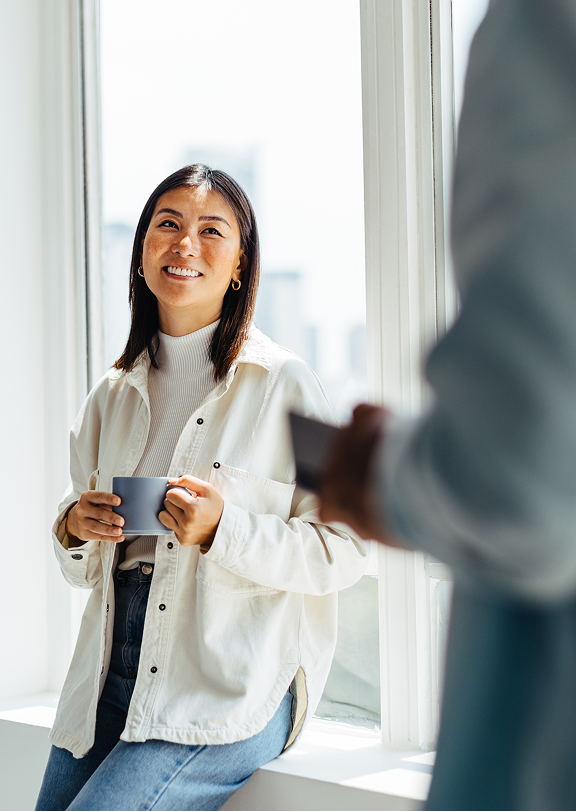 Kvinne som smiler mens hun holder en kaffekopp og ser på en annen person innendørs ved et vindu i dagslys.