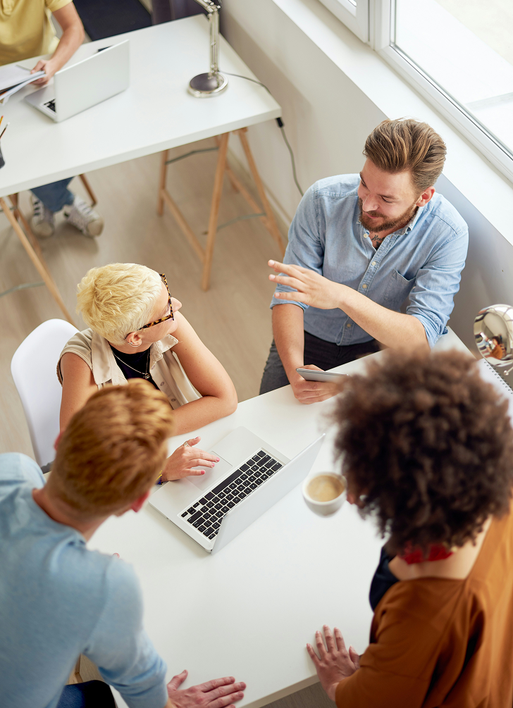 Fire personer som sitter rundt et hvitt bord med en bærbar PC, og diskuterer i et lyst kontor nær et vindu.