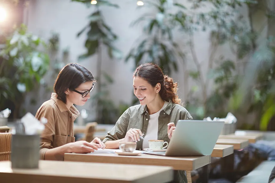To kvinner sitter ved et bord med en laptop og kopper, og samtaler i en lys kafé med planter i bakgrunnen.