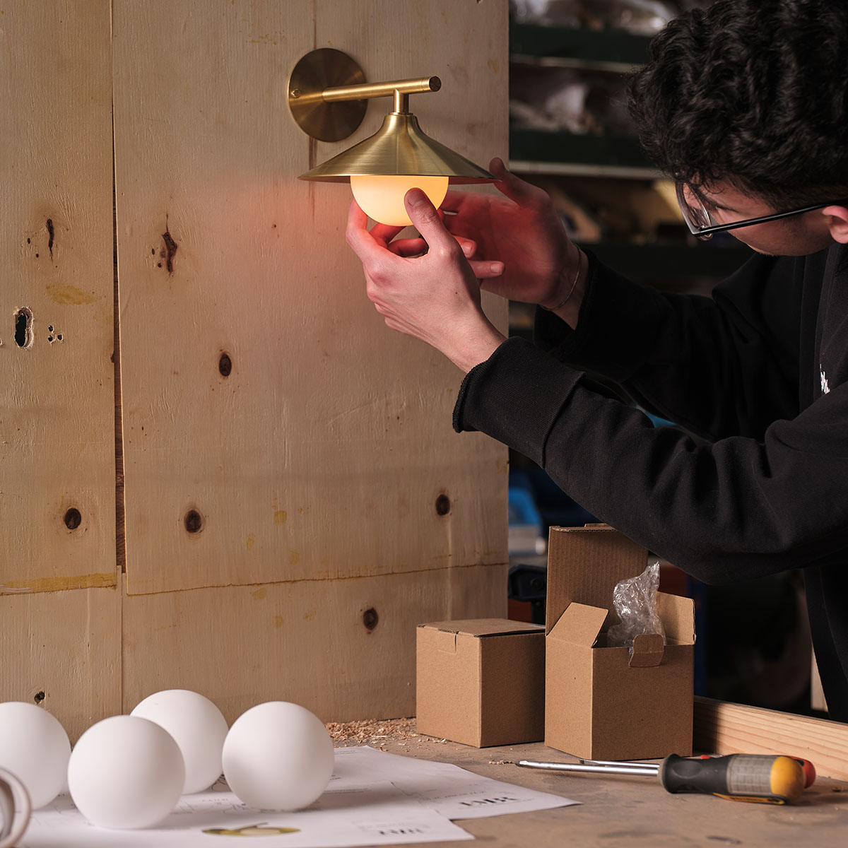 Craftsperson adjusting a brass wall light mounted on a workshop panel.