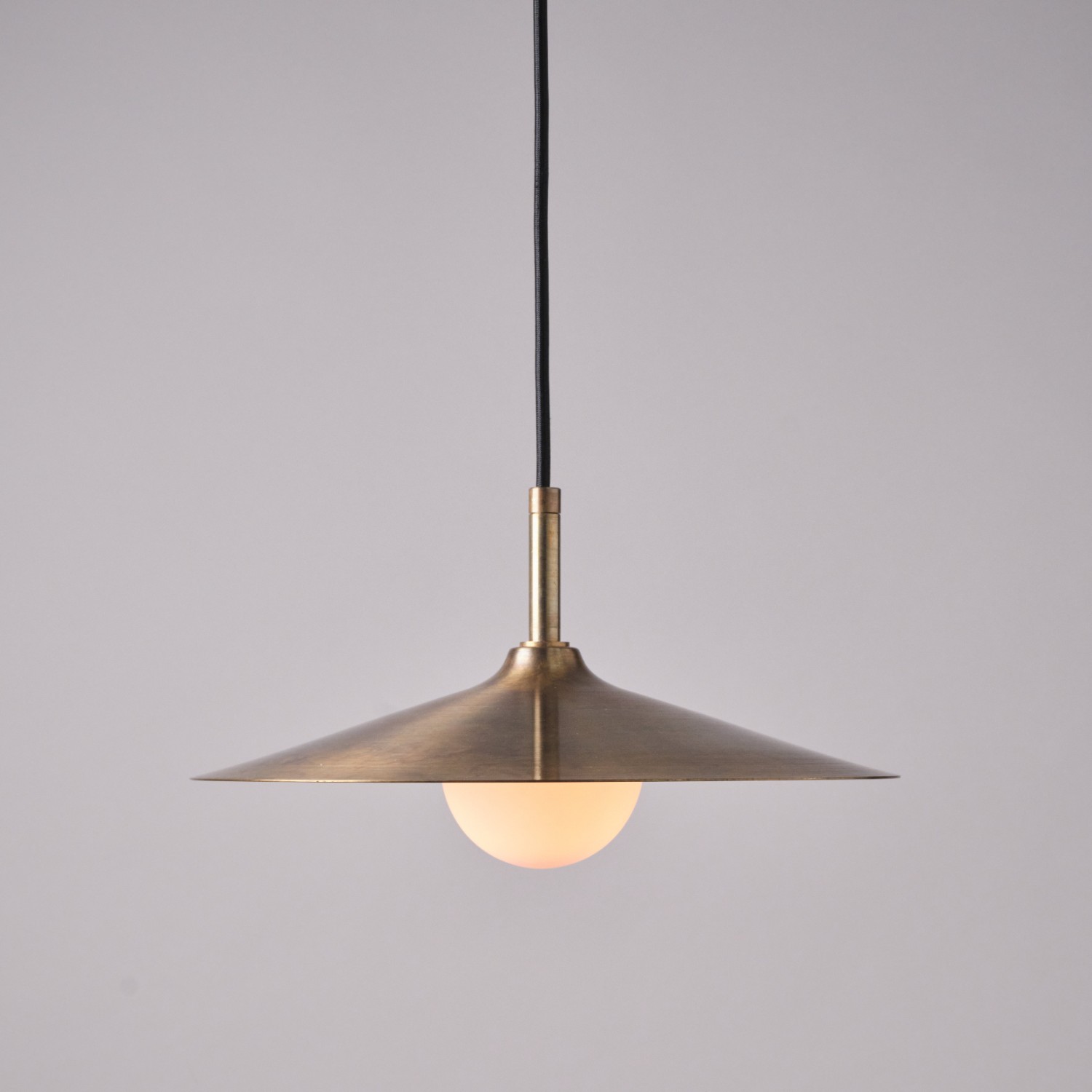 Large antique brass pendant light with a shallow conical shade and glowing globe bulb.