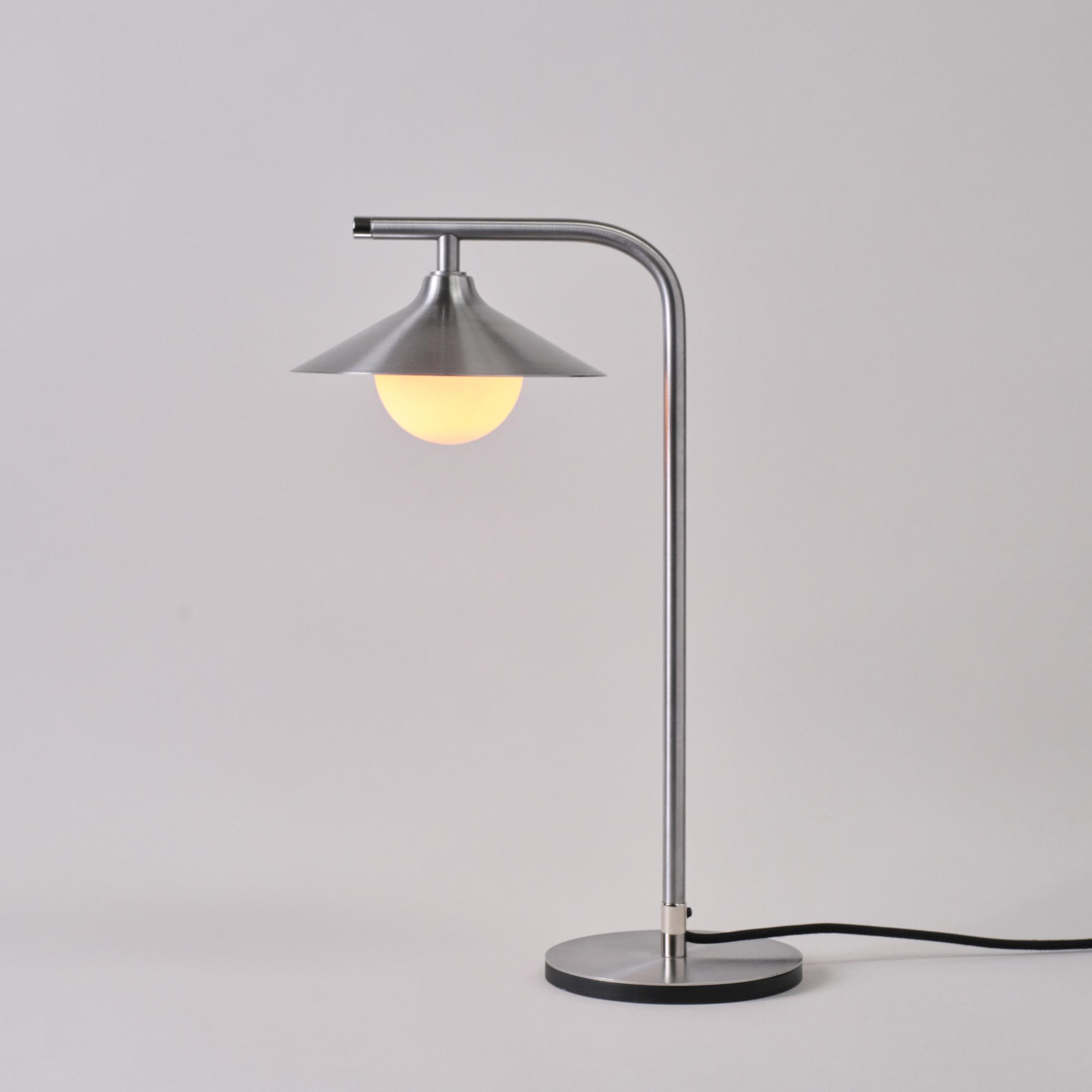 Mettle Alba table lamp in mild steel with a curved stem and glowing globe bulb.