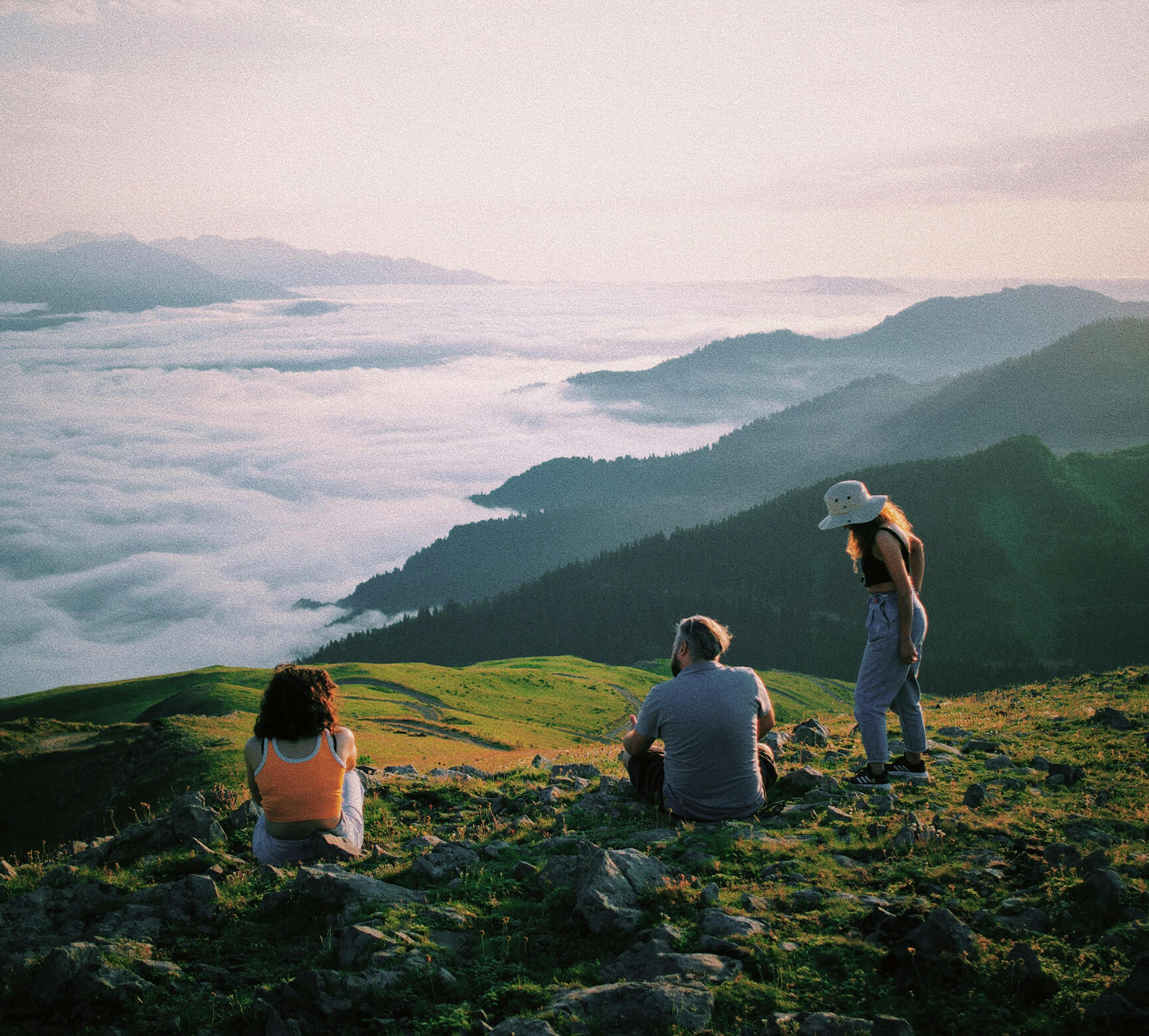 Three people enjoying a scenic view on a grassy mountain overlooking fog-covered valleys and distant hills at sunrise or sunset.