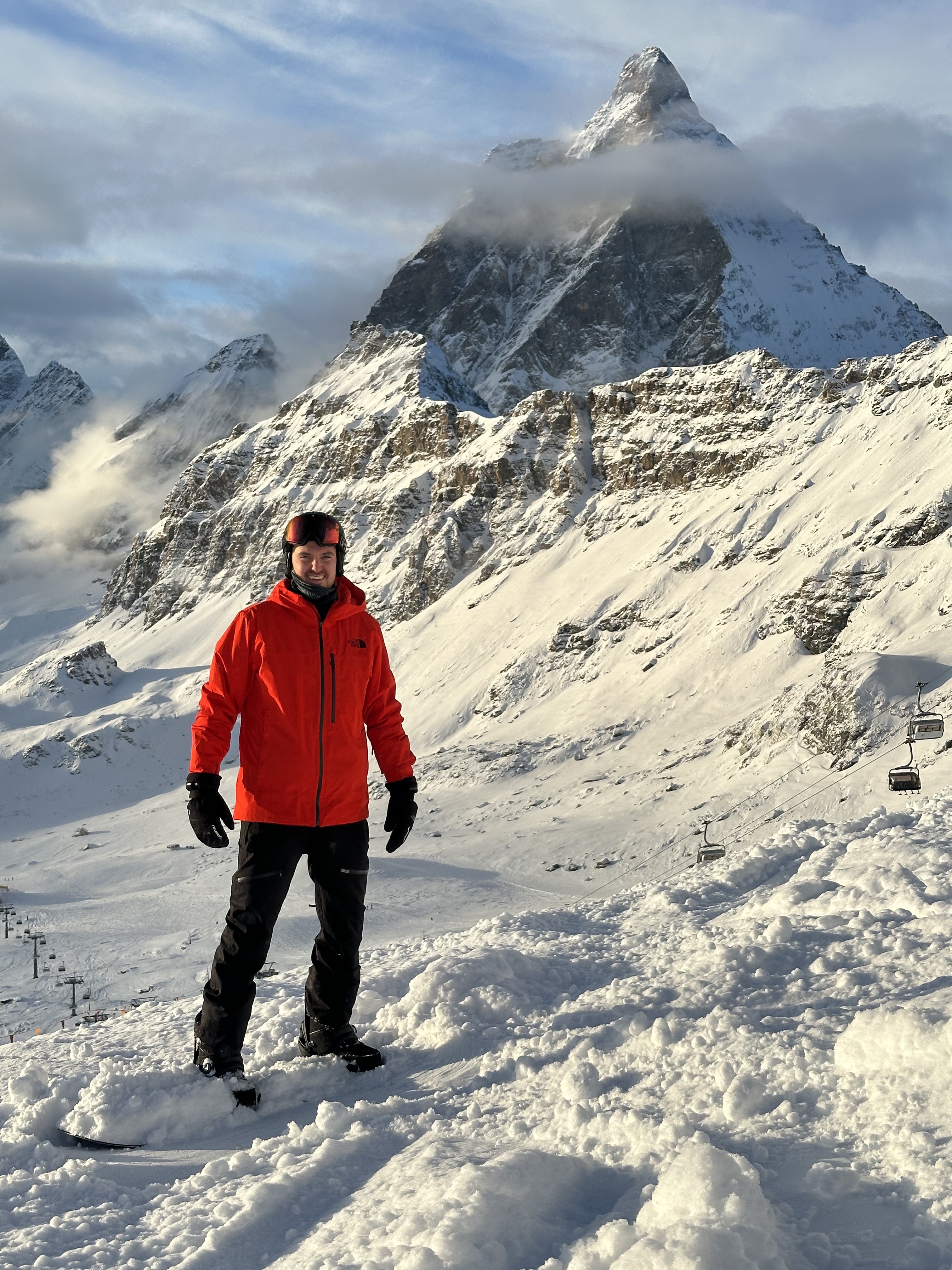 Smiling man in red jacket and black ski pants snowboarding in snowy mountain landscape with chairlift and a tall peak in the background.