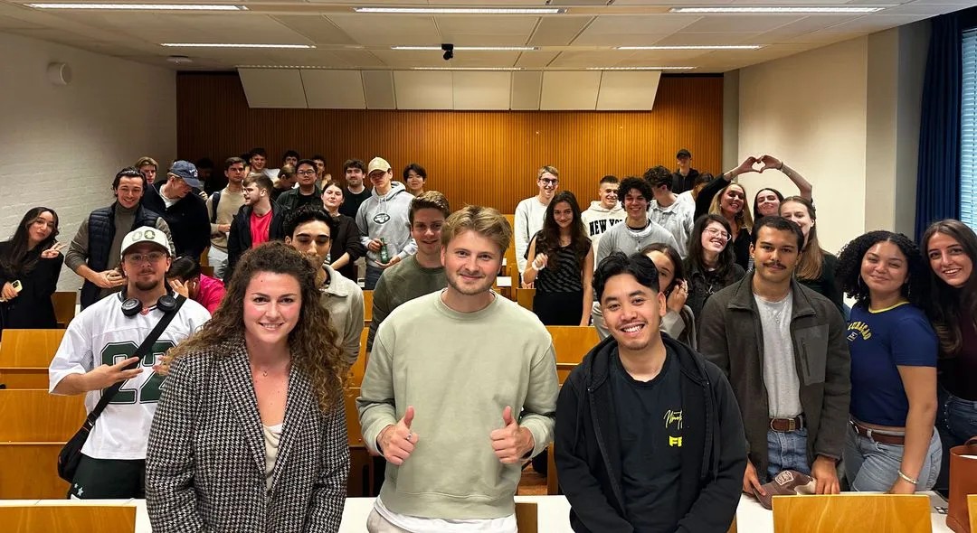 Group of young adults smiling and posing in a classroom with rows of wooden desks and a wood-paneled wall.