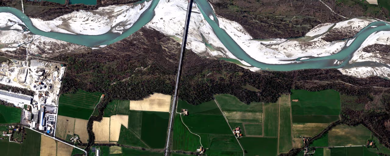 Aerial view showing a winding light blue river flowing through green and tan farmland with scattered buildings and wooded areas.
