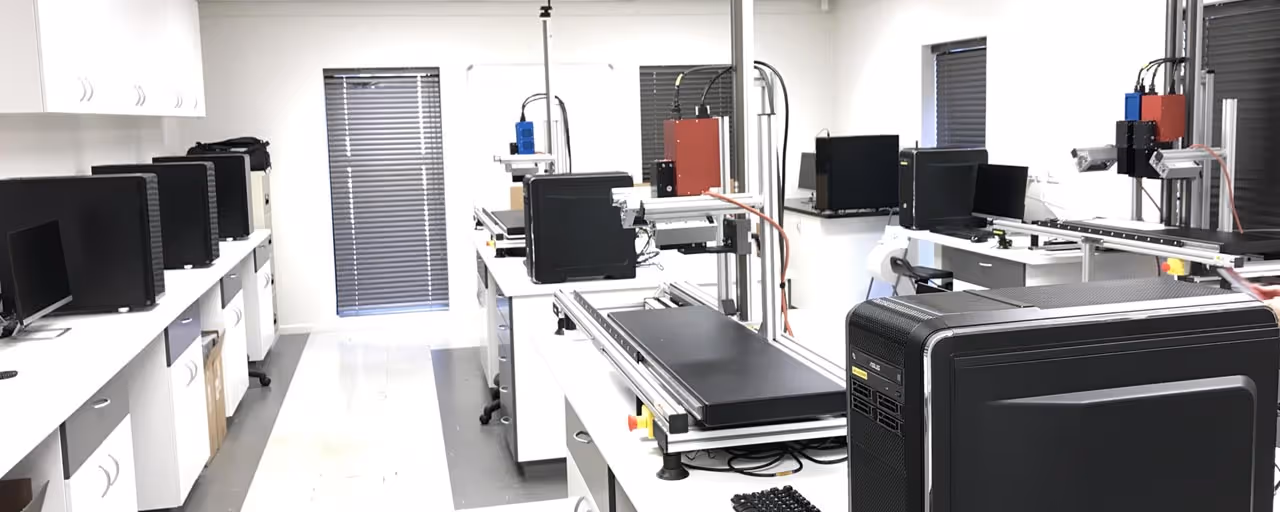 Modern laboratory room with multiple desktop workstations and specialized scanning or measuring equipment on white benches.