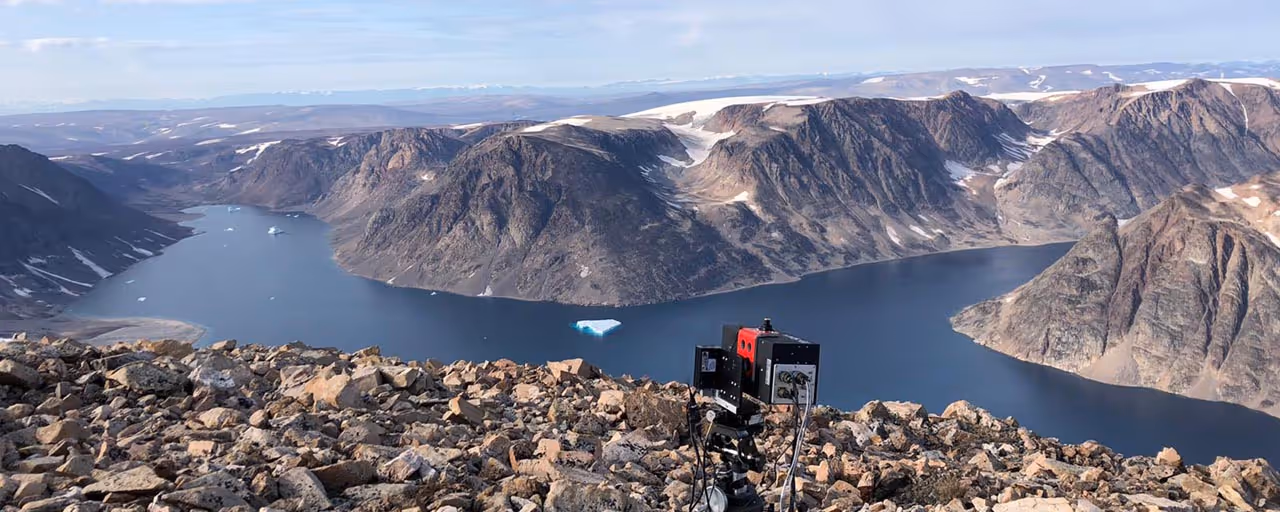 Scientific camera equipment set up on rocky terrain overlooking a fjord with dark blue water and surrounding snow-capped mountains.