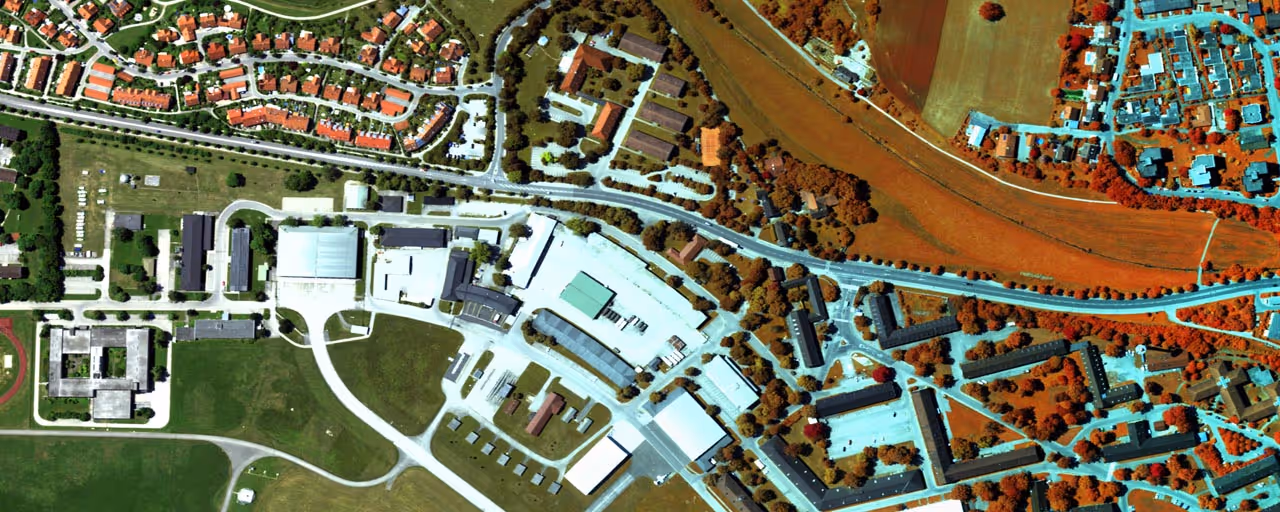 Aerial view of a suburban area with residential houses, industrial buildings, roads, and green fields with trees appearing in orange hues, indicating a false-color infrared image.