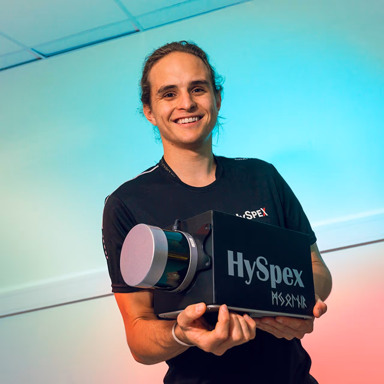 Smiling man holding a black device labeled HySpex against a gradient blue and pink background.