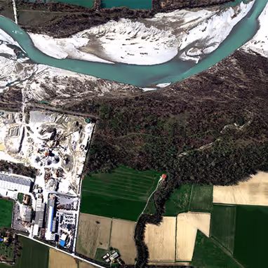 Aerial view showing a winding river with sandy banks, adjacent forested area, agricultural fields, and an industrial zone with buildings and machinery.