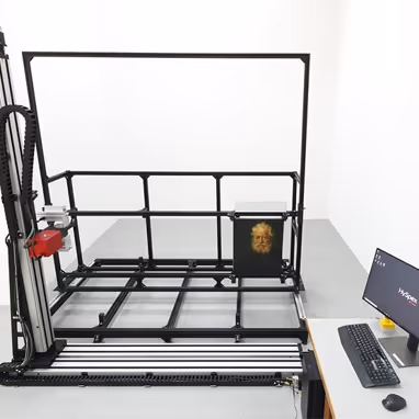 Large black metal frame structure with a vertical robotic arm and a painting of an elderly man displayed on the lower right part of the frame, next to a computer setup on a desk.