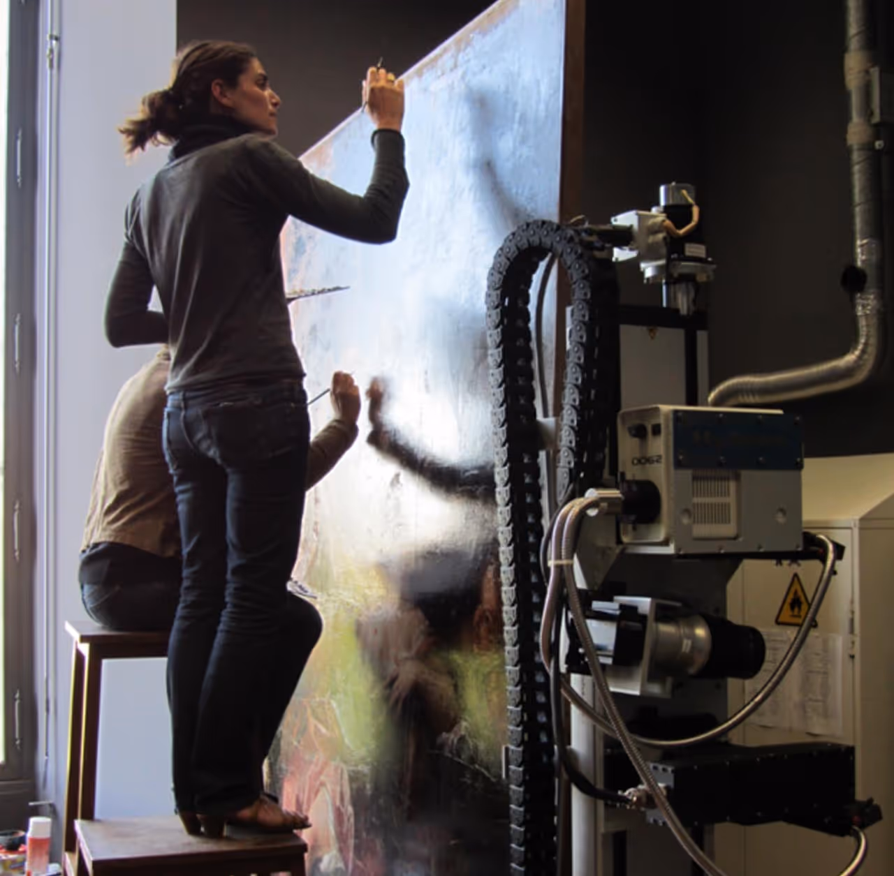 Two artists working on a large vertical canvas, one standing on a platform and painting, the other seated behind.
