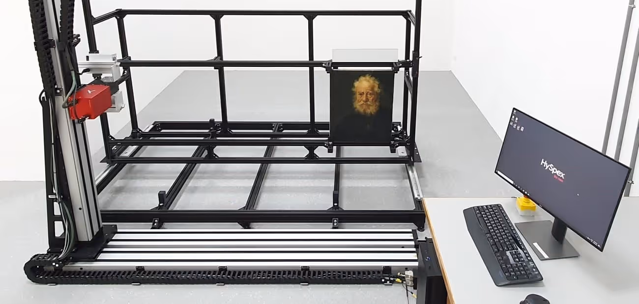 Art scanning device with a portrait of an elderly man attached to a black metal frame, next to a desk with a monitor displaying 'HySpex' and a keyboard.