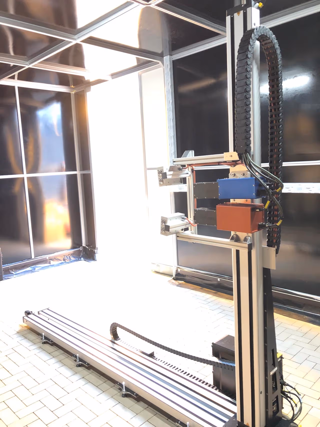 Tall vertical robotic arm assembly with cables, mounted on a long horizontal base inside a room with tiled floor and black walls.
