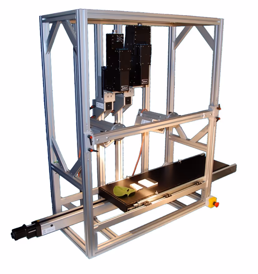 Laboratory rack system with two black sensors mounted above a conveyor belt holding a green leaf and two white objects.