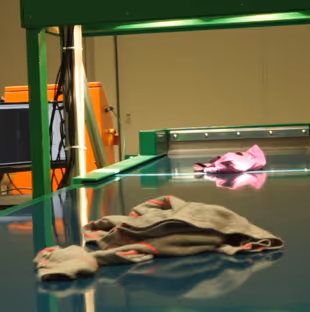 Clothes, including socks and a shirt, lying on a green conveyor belt in an industrial textile sorting facility.