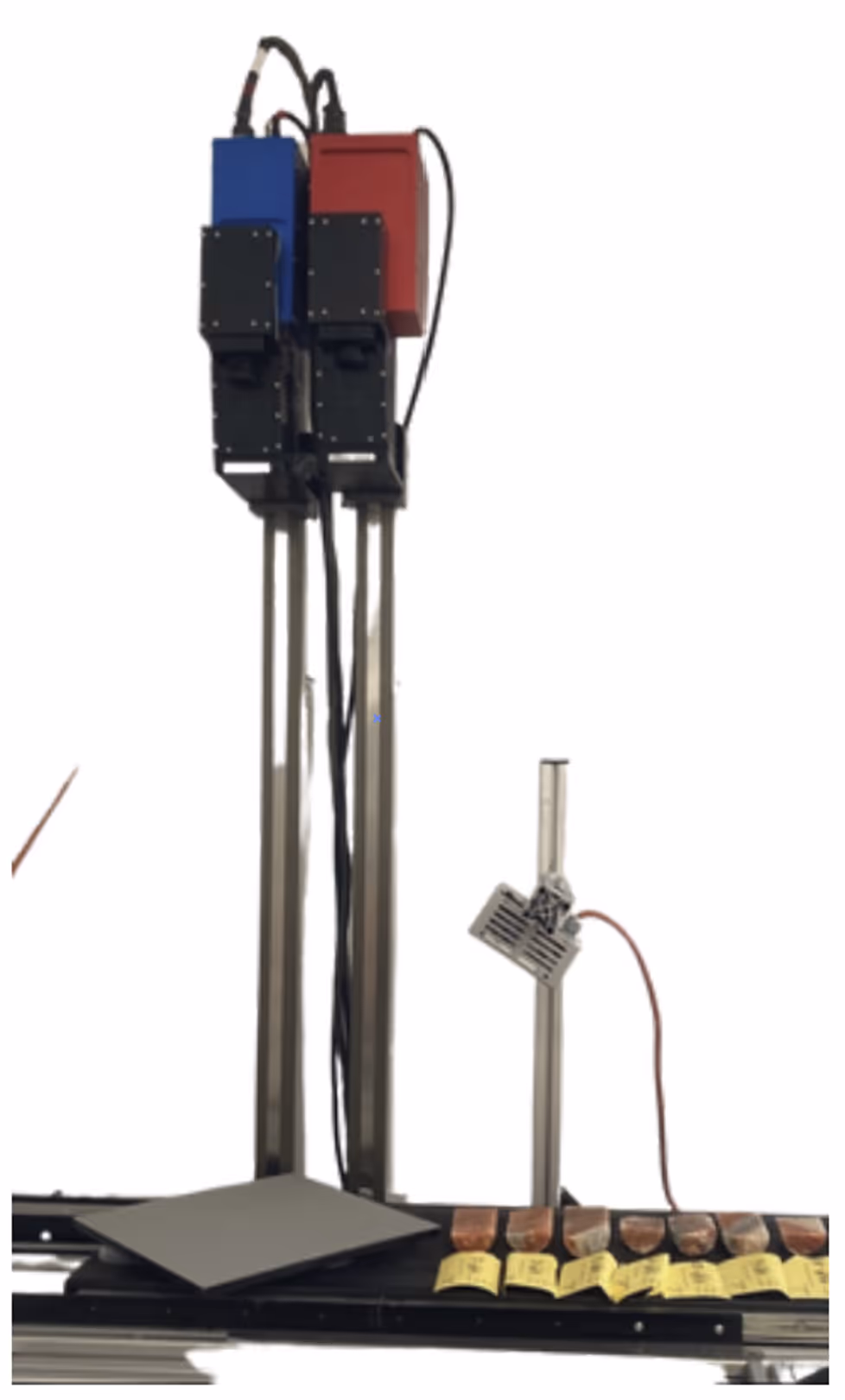 Scientific setup with two vertical metal stands holding blue and red box-like devices, a black rectangular panel laying flat, and a row of sample pieces labeled with yellow tags on a base platform.