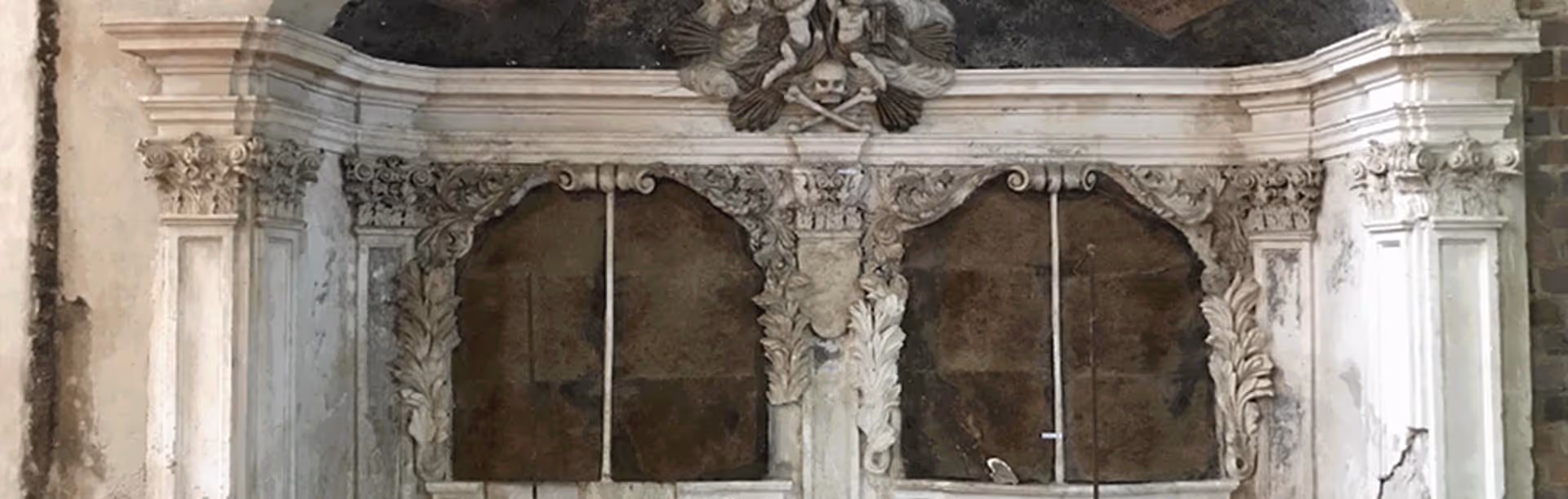 Ornate, weathered stone or marble altar with two arched recesses and a decorative skull and crossbones carving at the top center.