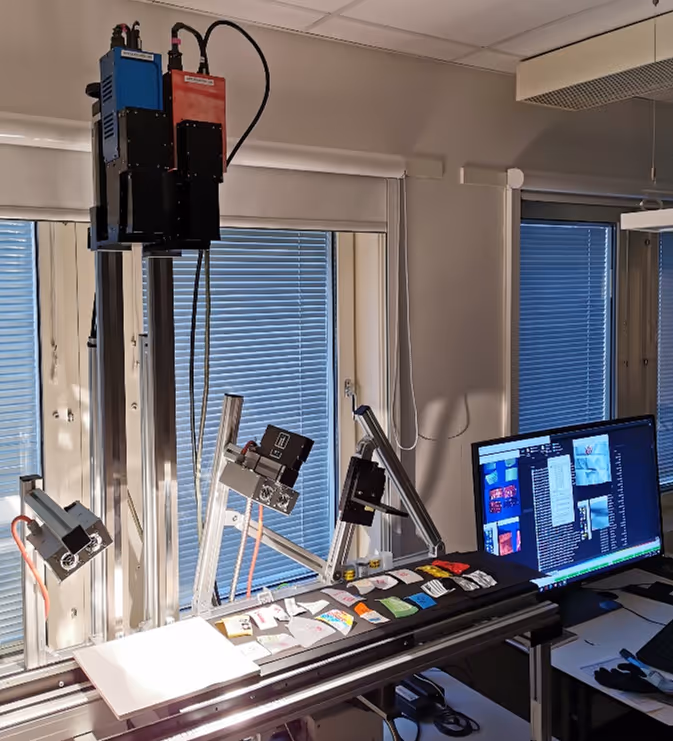 Laboratory setup with scanning equipment and camera system over a conveyor belt displaying various packets, connected to a computer screen showing data and images.