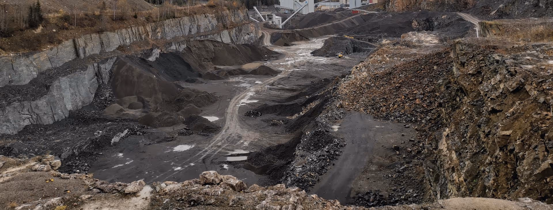 Large open-pit mine with exposed rock walls, dark soil piles, and a dirt road running through the center.