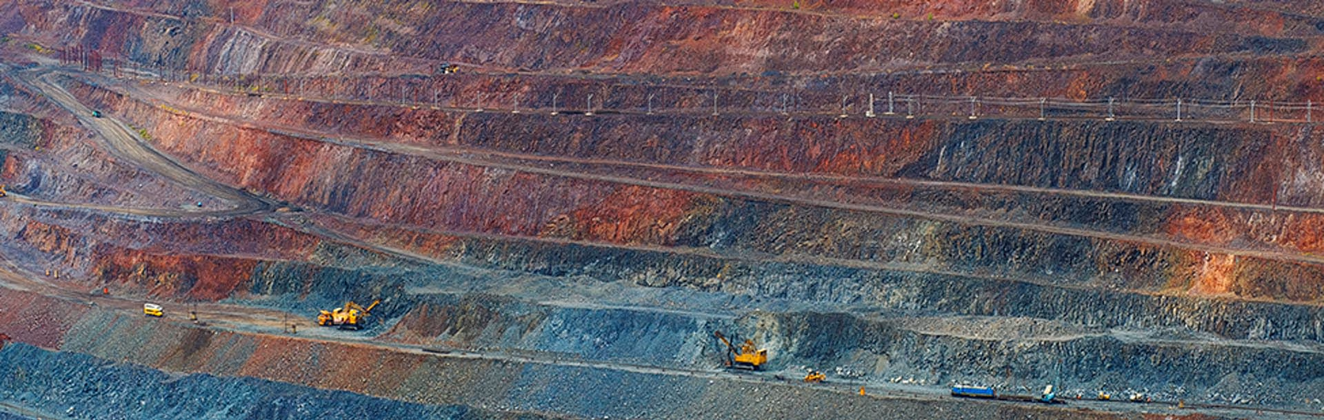Wide view of a terraced open-pit mine with layered rock formations in shades of red, brown, and gray and several yellow mining vehicles.