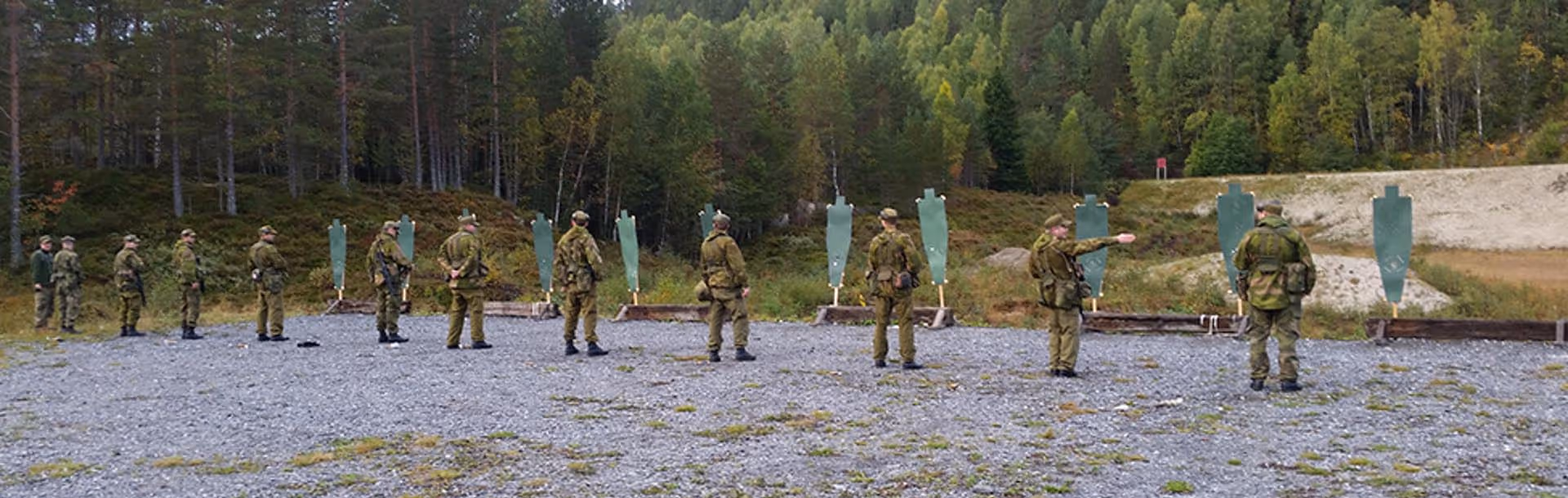 Group of soldiers in camouflage uniforms standing on a gravel area facing green shooting targets with a forest background.